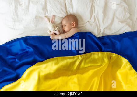 Ein schlafend Baby mit einem Lieblings-Plüschtier in der Hand, von oben mit der blau-gelben Flagge der Ukraine bedeckt. Sorgloser Schlaf eines Babys mit einem weichen zu Stockfoto