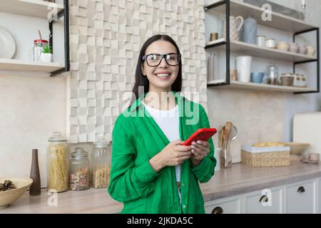 Junge schöne Frau in Brille und grünem Hemd in der Küche mit einem roten Telefon, lächelnd und Blick auf die Kamera, Porträt einer glücklichen Blondine Stockfoto