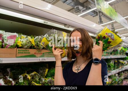 Moskau, Russland, 2020. August: Eine junge Frau in einer lustigen Schutzmaske mit einem Hundegesicht hält Grünflächen in einem Lebensmittelgeschäft. Stockfoto