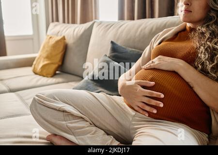 Hände einer jungen Schwangeren in braunem Tanktop und grauer Jogginghose sitzen auf einer bequemen Couch mit Kissen im Wohnzimmer Stockfoto