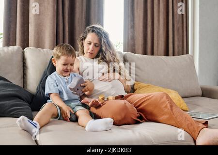 Junge Schwangere in Casualwear sitzen auf weicher bequemer Couch neben ihrem niedlichen kleinen Sohn, während beide mit Spielzeug spielen Stockfoto