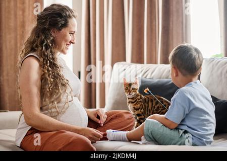 Glückliche junge Schwangere, die ihren niedlichen kleinen Sohn ansieht, der mit einer Katze aus bengalen spielt, während sie sich auf der Couch im Wohnzimmer entspannt Stockfoto