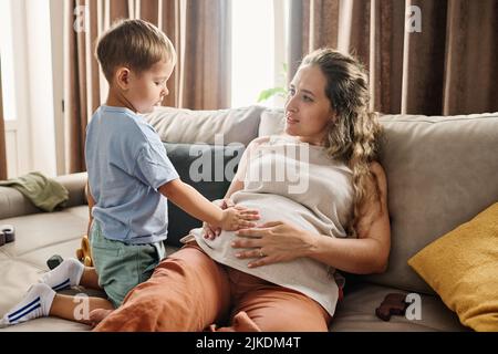 Neugieriges Kleinkind, das die Hand am Bauch seiner Schwangeren auf dem Sofa sitzend hält, während es Freizeit mit ihrem niedlichen kleinen Sohn zu Hause verbringt Stockfoto