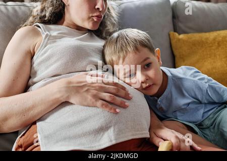 Neugieriger kleiner Junge, der seinen Kopf auf dem Bauch seiner schwanger Mutter hält, während er dem Herzschlag des Mutterleibes lauscht, während er auf der Couch sitzt Stockfoto