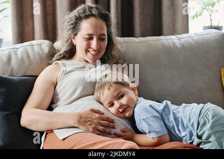 Happy adorable Kleinkind Junge hören Herzschlag des Schlages des Unten im Bauch seiner Schwangeren Mutter, während das Ohr auf ihrem Bauch halten Stockfoto