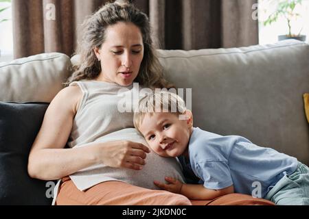 Niedlicher kleiner Junge in Casualwear, der das Ohr nahe am Bauch seiner Schwangeren hält, während er versucht, den Herzschlag eines zukünftigen Babys zu fangen Stockfoto