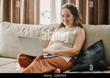 Glückliche junge, schwanger Geschäftsfrau oder freiberufliche Freiberuflerin, die während des Netzwerkes auf dem Laptop-Tastenfeld tippt, während sie auf den Bildschirm schaut Stockfoto