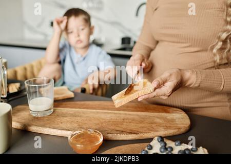 Hände einer jungen Schwangeren, die Honig oder Marmelade auf einer Scheibe Weizenbrot verteilt, während sie einen Snack für ihren kleinen Sohn zum Frühstück zubereitet Stockfoto