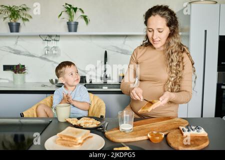 Junge Schwangere, die am Küchentisch steht und Honig oder Marmelade auf einer Weizenbrotscheibe verteilt, während sie das Frühstück zubereitet Stockfoto