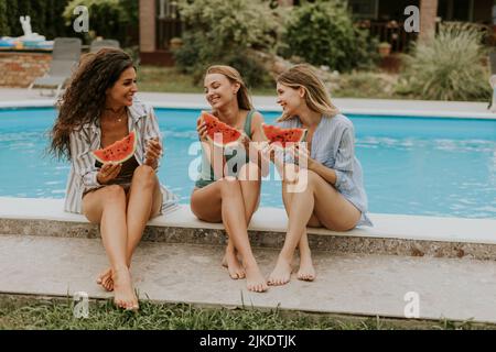 Drei niedliche junge Frauen sitzen am Pool und essen Wassermellon im Hinterhof des Hauses Stockfoto