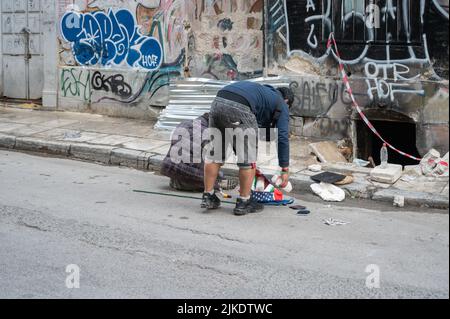 ATHEN, GRIECHENLAND - 14. MAI 2022: Ein Obdachloser schläft auf einem Bürgersteig in der Innenstadt von Athen. Touristen kommen vorbei. Stockfoto