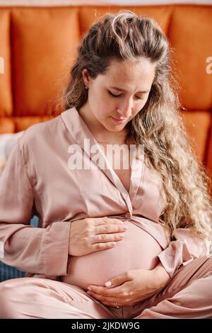 Eine Schwangere im pinken Seiden-Pyjama blickt auf ihren Bauch und berührt ihn, während sie auf dem Bett sitzt und mit ihrem zukünftigen Baby kommuniziert Stockfoto