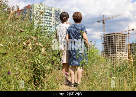 Zwei Frauen stehen auf grünem Rasen und schauen sich die neuen Häuser von Wohnbezirks- und Baukranen an. Käufer, die eine Wohnung wählen Stockfoto