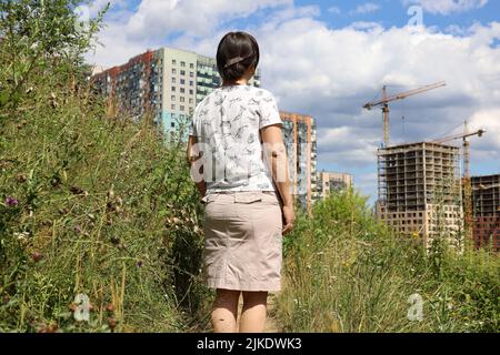Frau, die auf grünem Rasen steht und sich die neuen Häuser von Wohnbezirks- und Baukranen anschaut. Käufer, die eine Wohnung wählen Stockfoto