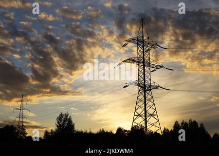 Silhouetten von Hochspannungstürmen mit elektrischen Drähten auf dem Hintergrund des Sonnenuntergangs Himmel und dunklen Wolken. Stromübertragungsleitungen im Abendwald Stockfoto