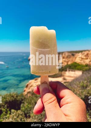 Hand hält ein Eis mit blauem Meer und Klippen im Hintergrund. Stockfoto