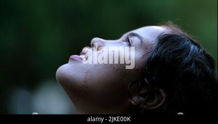 Junge Frau, die draußen im Regen steht, Mädchen, die die Augen zum Himmel öffnen und Hoffnung in einem regnerischen Tag fühlt Stockfoto