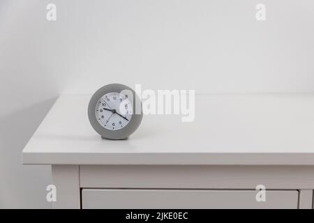 Eine runde graue Uhr zeigt zwanzig nach neun auf einer weißen Kommode. Stockfoto