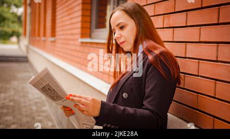 Junge Frau liest Zeitung auf der Straße. Seitenansicht einer jungen Frau mit langen Haaren in legerer Kleidung, die auf der Straße steht und Zeitung liest. Stockfoto