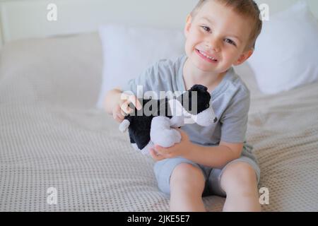 Nahaufnahme eines kleinen glücklichen kaukasischen Jungen in einem grauen T-Shirt liegt mit einem Spielzeughund auf dem Bett Stockfoto