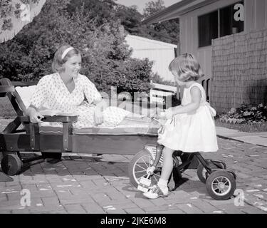 1960S LÄCHELNDE FRAU MUTTER AUF HINTERHOF BACKSTEIN TERRASSE AUF CHAISELONGUE LIEGEND BUCH VON KLEINEN MÄDCHEN TOCHTER REITEN DREIRAD ZU LESEN - J11724 HAR001 HARS ALTE ZEIT NOSTALGIE ALTE MODE 1 FITNESS JUGENDLICH GESUNDES GLEICHGEWICHT FAMILIEN LIFESTYLE CHAISE PARENTING BRICK WEIBCHEN TERRASSE RADFAHREN HEIM LEBEN KOPIE RAUM HALBE LÄNGE DAMEN LOUNGE TÖCHTER PERSONEN VERTRAUEN FAHRRÄDER TRANSPORT B & W FAHRRÄDER AKTIVITÄT GLÜCK KÖRPERLICHE DREIRAD FREIZEIT KRAFT UND STOLZ AUF FLEXIBILITÄT MUSKELN UNTERSTÜTZEN WACHSTUM JUGENDLICHE MITTLERE ERWACHSENE MITTLERE ERWACHSENE FRAU MÜTTER ENTSPANNUNG ZWEISAMKEIT TRIKE SCHWARZ UND WEISS KAUKASISCHE ETHNIE Stockfoto