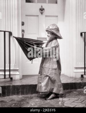 1920S KLEINES MÄDCHEN IM REGEN TRÄGT EINEN LANGEN REGENMANTEL UND HAT SOU’WESTER FOUL WEATHER GEAR UND SETZT EIN REGENSCHIRM - J1280 HAR001 HARS GEWINNEN HEIMLEBEN LANG REGNERISCHE WOHNANLAGE AUSSCHNITT REGNET GEBÄUDE B&W FRÖHLICHES ABENTEUER UND AUFREGUNG AUF HÄUSER LÄCHELT FRÖHLICH WOHNSITZ VOR DER TÜR FOUL ANGENEHM ANGENEHM CHARMANT FOUL WETTER JUGENDLICHE LIEBENSWERT LIEBENSWERT ENTZÜCKEND ANSPRECHEND SCHWARZ UND WEISS KAUKASISCHE ETHNIZITÄT HAR001 ALTMODISCH Stockfoto