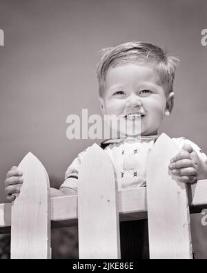 1950S PORTRAIT KLEINKIND JUNGE LÄCHELND STEHEND AM WEISSEN PFOSTENZAUN BLICK AUF DIE KAMERA - J5411 HAR001 HARS GESUNDHEIT ZU HAUSE LEBEN KOPIEREN RAUM MÄNNER VERTRAUEN S & W BLICKKONTAKT GLÜCK KOPF UND SCHULTERN FRÖHLICH FÜHRUNG NIEDRIG WINKEL SCHÖNER STOLZ LÄCHELT FREUDVOLLES BABY JUNGE ANGENEHM ANGENEHM CHARMANT EIFRIG WACHSTUM JUVENILES LIEBENSWERT LIEBENSWERT ANSPRECHEND SCHWARZ UND WEISS KAUKASISCHEN ETHNIE HAR001 ALTMODISCH Stockfoto