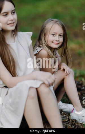 Zwei kleine Mädchen in Sommerkleidern sitzen auf einem Stumpf im Park. Ein Mädchen schaut in die Kamera Stockfoto