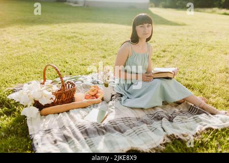 Eine schöne junge Europäerin sitzt auf einer Decke und hält ein Buch zum Lesen. Ein Mädchen auf einem Picknick in einem Sommerpark. Stockfoto