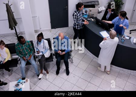 Draufsicht auf den Wartebereich des Krankenhauses, wo ein leitender Arzt asiatische Patienten an der Rezeption in die Klinik einweihend. afroamerikanischer Arzt füllt Formular für Patiententermin in medizinischen Einrichtungen aus. Stockfoto