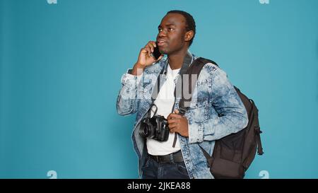 afroamerikanischer Fotograf im Gespräch mit dem Smartphone, Vorbereitung auf Urlaub Reise mit Rucksack und nehmen professionelle Bilder. Ferngesteuerte Unterhaltung vor der Aufnahme von urbane Abenteuerfotos. Stockfoto