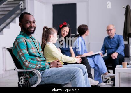 Porträt eines afroamerikanischen Patienten, der die Kamera anlächelt, während eine professionelle Krankenschwester einen älteren Mann in die Klinik einlässt. Selbstbewusster Mann, der sich mit verschiedenen Menschen in der Krankenhausaufnahme niedergelassen hat. Stockfoto