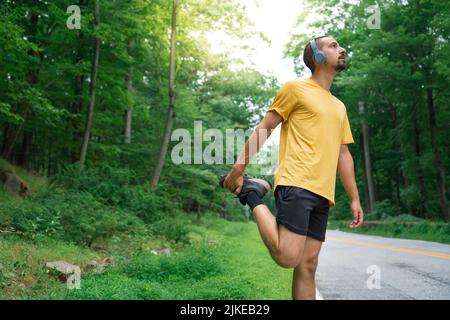Junger Fitnessmann, der sich die Beine streckt, bevor er mit Ohrhörern auf der Straße in der Natur läuft. Gesunder Lebensstil und Sportkonzepte Stockfoto