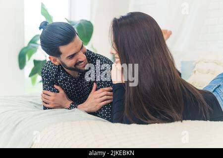 Dunkelhaariger Mann mit einem Bart, der seine Freundin lächelnd anschaut und sich morgens auf einem Bett entspannt. Junges Paar. Innenaufnahme. Hochwertige Fotos Stockfoto