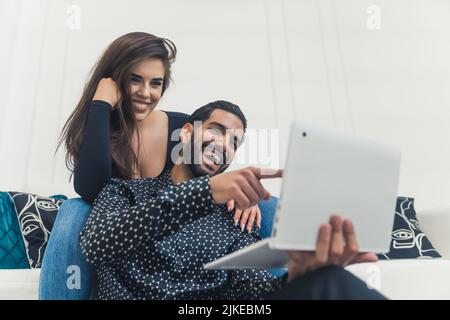 Dunkelhaariger Mann mit lachendem Bart und dem Hinweis auf den Laptop, wobei seine Freundin lächelnd auf der Couch saß und sich auf ihn lehnte. Innenaufnahme. Hochwertige Fotos Stockfoto