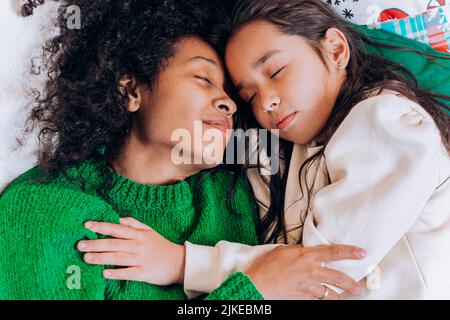 Afroamerikanische Mutter mit lockigen Haaren umarmt die brünette Tochter und lacht, während sie an Weihnachten gemütlich im Wohnzimmer auf Kissen liegt Stockfoto