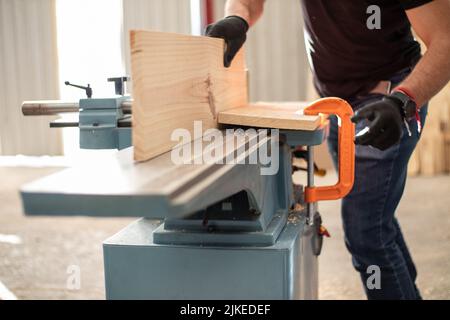 Junger Zimmermann, der an einer elektrischen Tischlermaschine arbeitet, schneidet in einer Schreinerei mit schwarzen Sicherheitshandschuhen ein Holzbrett Stockfoto