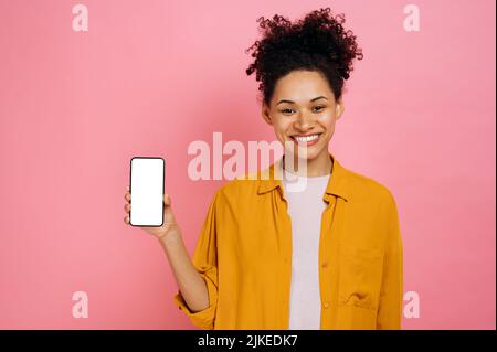 Positive schöne afroamerikanische junge Frau mit lockigem Haar, Mobiltelefon mit leerem weißen Bildschirm in der Hand haltend, Kopierraum für Werbung, freundlich lächelnd, stehend über einem isolierten rosa Hintergrund Stockfoto