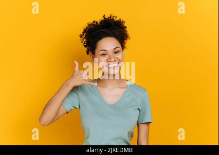 Rufen Sie mich an. Positives afroamerikanisches Millennial-Mädchen, das die Hand am Ohr hält, mit den Fingern ein Telefonschild zeigt, nach der Nummer fragt, auf einem isolierten orangefarbenen Hintergrund steht, lächelnd auf die Kamera blickt Stockfoto