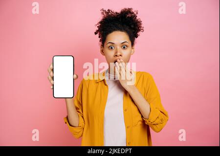 Verwirrt schockiert afroamerikanische lockige junge Frau in Freizeitkleidung, zeigt Smartphone mit weißem Mockup-Bildschirm, auf isoliertem rosa Hintergrund stehend, sieht überrascht Kamera, bedeckt ihren Mund Stockfoto