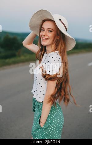 Eine rothaarige Frau mit Sommersprossen, die einen Hut trägt, allein auf dem Land posiert, lächelt und die Kamera anschaut. Sommerwetter, grünes Feld. Attraktiv Stockfoto