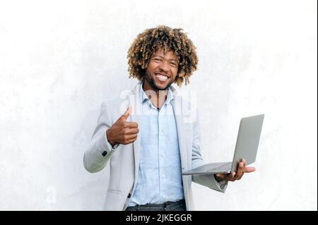 Stolzer erfolgreicher, selbstbewusster afroamerikanischer oder brasilianischer Mann, elegant gekleidet, mit offenem Laptop in der Hand, steht auf isoliertem Hintergrund, blickt lächelnd auf die Kamera, zeigt Daumen nach oben Stockfoto