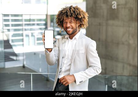 Erfolgreicher, freundlicher afroamerikanischer oder brasilianischer lockiger Mann, steht im Freien in stilvoller Kleidung, zeigt Smartphone mit weißem Mockup-Bildschirm, Platz für Werbung, schaut auf die Kamera, lächelt Stockfoto