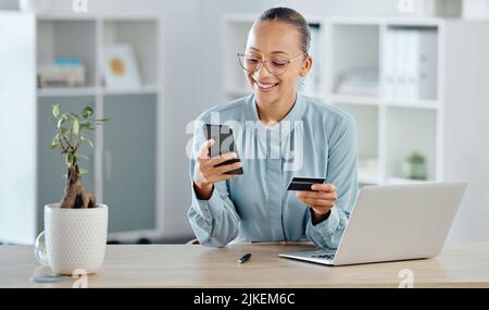 Weibliche Führungskraft online einkaufen, mit einer Corporate Kreditkarte auf einem Telefon kaufen. Eine lächelnde Frau, die Geschäftsrechnungen bezahlt, sitzt am Schreibtisch mit Stockfoto