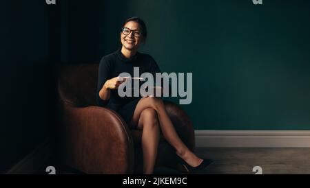 Lächelnde Frau, die auf einem Stuhl sitzt und die Kaffeetasse hält. Glückliche asiatische Frau, die Kaffee genießt, während sie auf einem Sessel sitzt. Stockfoto