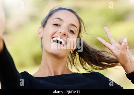 Frieden im aus. Beschnittenes Porträt einer attraktiven und athletischen jungen Frau, die im Freien steht und den Frieden gestikelt. Stockfoto