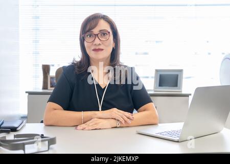 Porträt einer brünette Latina-Ärztin, die in ihrem Büro neben ihrem Laptop auf die Kamera schaut Stockfoto