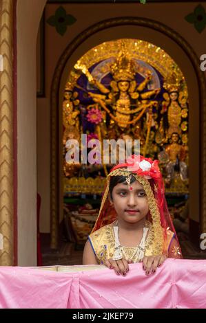 Howrah, Indien -26.. Oktober 2020 : Bengalisches Mädchen in festlichem Kleid, lächelnd und posiert mit Göttin Durga im Hintergrund, innen dekoriert Haus. Stockfoto