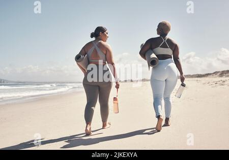 Wir haben es uns zur Priorität gemacht, regelmäßig Yoga zu praktizieren. Zwei junge Frauen, die mit ihren Yogamatten am Strand spazieren. Stockfoto