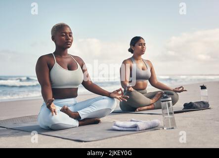 Ich fühle mich in Frieden, wenn ich um dich herum bin. Zwei junge Frauen meditieren während ihrer Yoga-Routine am Strand. Stockfoto